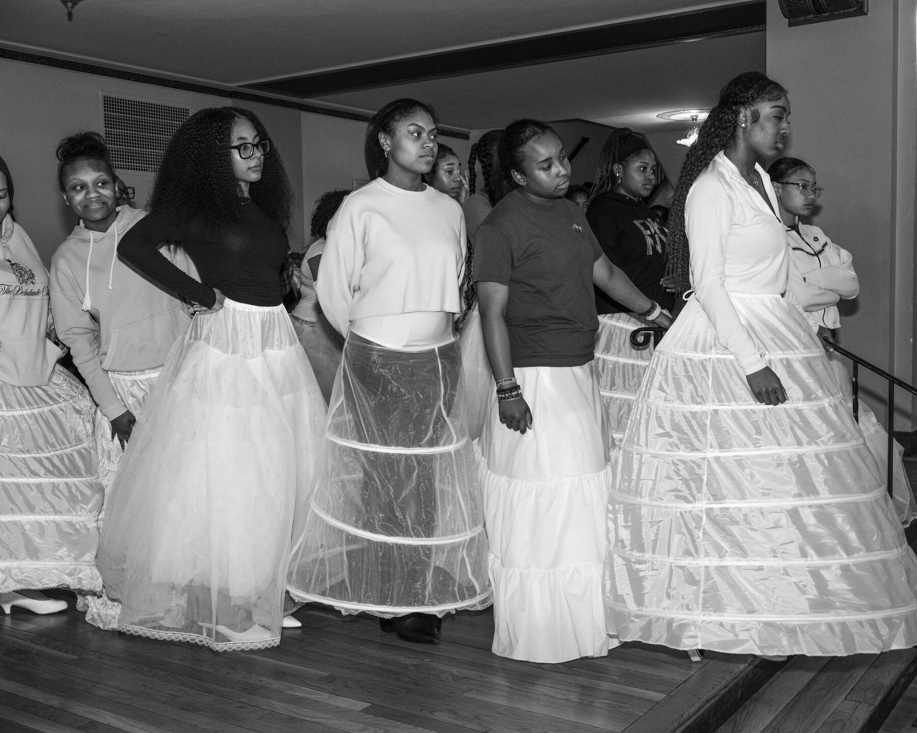 A group of young women wearing white hoop skirts or petticoats and T-shirts of sweatshirts. They stand at the top of a wood staircase, looking at something off camera to the right.