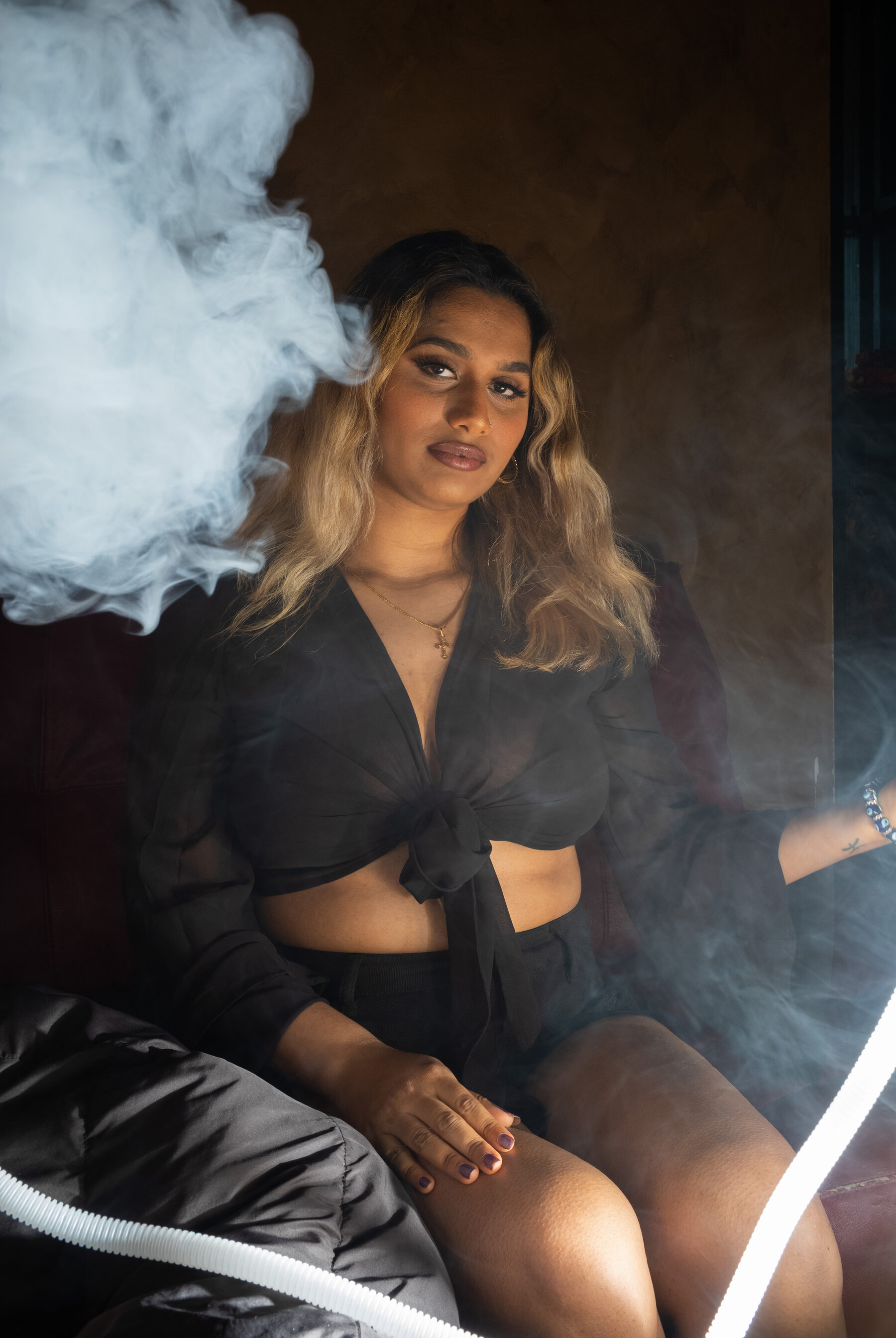 A young woman wearing a long-sleeved sheer black crop top with a tie front and black shorts and sitting in a hookah lounge. She holds the end of a long clear plastic hose, and a cloud of smoke floats next to her head.